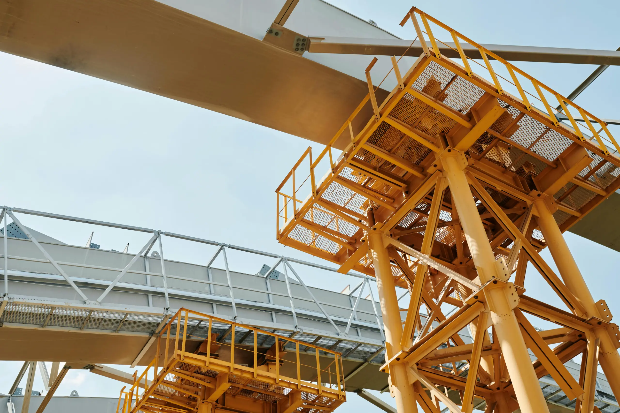 Yellow metal industrial conveyor system with walkways and supports against a bright sky.