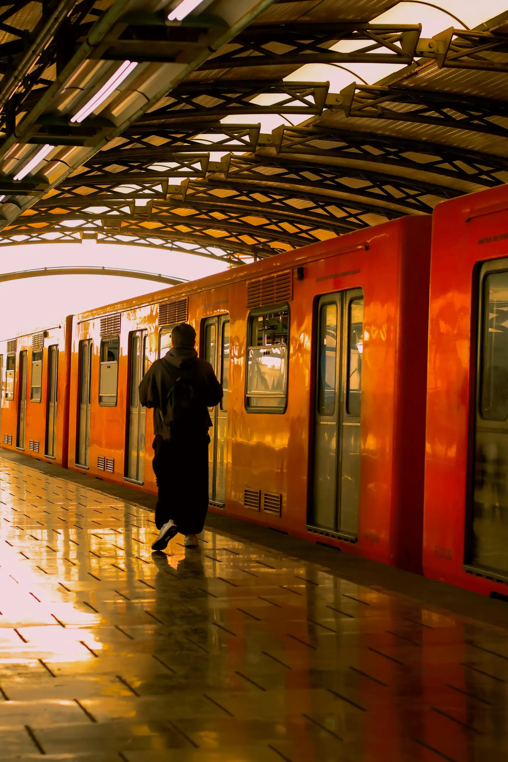 Person walks toward an orange subway train at a station with a patterned ceiling.